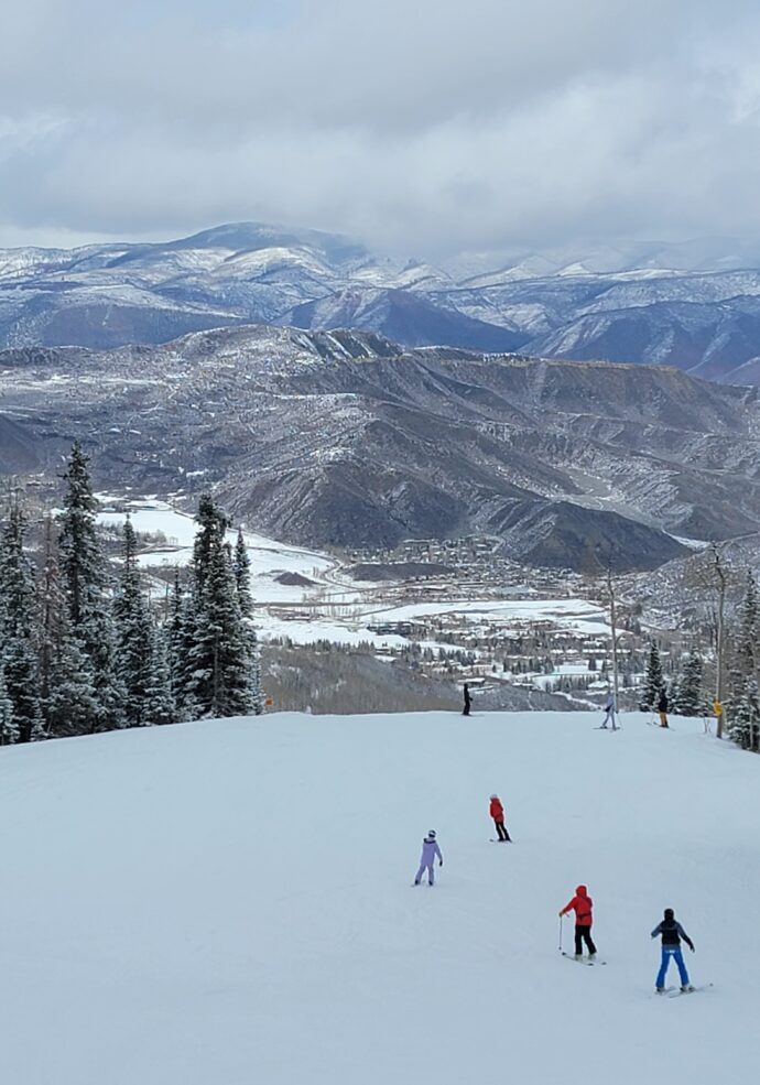A snowy view of skiers at Snowmass Ski Are with mountains in the distance